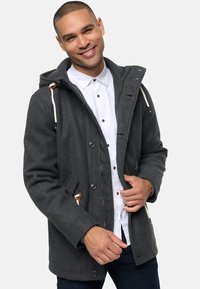 Grå uldblandingsfrakke med hætte, frontknapper og dobbelt sidelommer. Har snøringer og trækkelementer i træ. Stylet over en hvid skjorte.