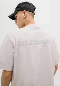 Šviesiai rožinė trumpomis rankovėmis marškinėliai su užrašu "GLENDALE" ir "Est. 1990, LOCAL MERCH" tekstūrinėmis pilkomis raidėmis ant nugaros. Nešiojama juoda kepurė.