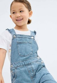 Jeans kombinezoni z nastavljivimi naramnicami, sprednjim žepom in srebrnim okovjem, oblečeni preko bele majice s pisanih cvetličnim motivom.