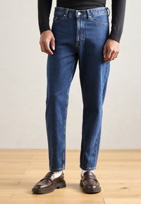 Denim jeans in dark blue with a high waist, straight-leg cut, subtle fading, and classic five-pocket design, paired with brown loafers.