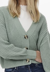 Lichtgroene gebreide cardigan met een V-hals, voorzien van twee grote bruine knopen en geribbelde manchetten, gedragen over een witte textuurtop.