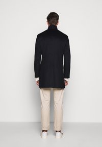 Cappotto di lana nero con colletto alto, maniche lunghe e vestibilità su misura, abbinato a pantaloni beige e sneakers bianche, visto da dietro.