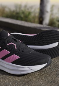 Czarne i różowe buty sportowe z białymi podeszwami spoczywające na chodniku, jeden but stoi pionowo, a drugi na boku ukazując wzór bieżnika.