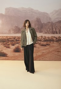 Brauner Hahnentrittmuster-Blazer mit doppelten Knöpfen, kombiniert mit einer cremefarbenen Bluse und schwarzen weiten Hosen. Neutraler Wüstenlandschaftshintergrund.