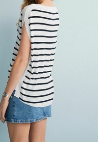 Kvinde iført en ærmeløs hvid skjorte med sorte vandrette striber og en blå denim miniskørt, stående op ad en lyseblå væg.