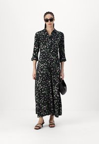 Robe noire à manches longues avec un motif floral en rose et vert. Dotée d'un devant à boutons, d'un col et d'une coupe décontractée. Portée avec des accessoires noirs.