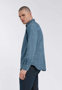 Camicia in denim blu chiaro con maniche lunghe, design con bottoni e due spacchi laterali in fondo. Il tessuto appare liscio con una vestibilità casual.