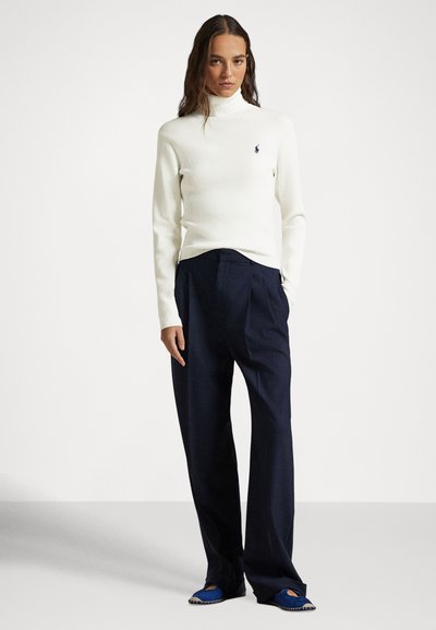 Žena oblečená v bielom roláku, voľných tmavomodrých nohaviciach a modrých espadrilkách, stojaca na jednoduchom pozadí.