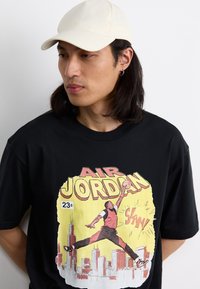 T-shirt noir avec un graphique coloré d'un joueur de basket-ball, entouré d'un paysage urbain, avec le texte "AIR JORDAN" et une casquette beige.