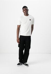 White short-sleeve t-shirt with a round black logo on the chest, paired with black loose-fit pants and black sneakers with white soles.