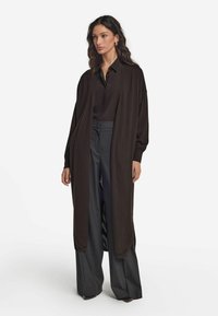 Lång brun kofta i mjukt tyg, med öppen fram och vida ärmar, tillsammans med mörka höga byxor.