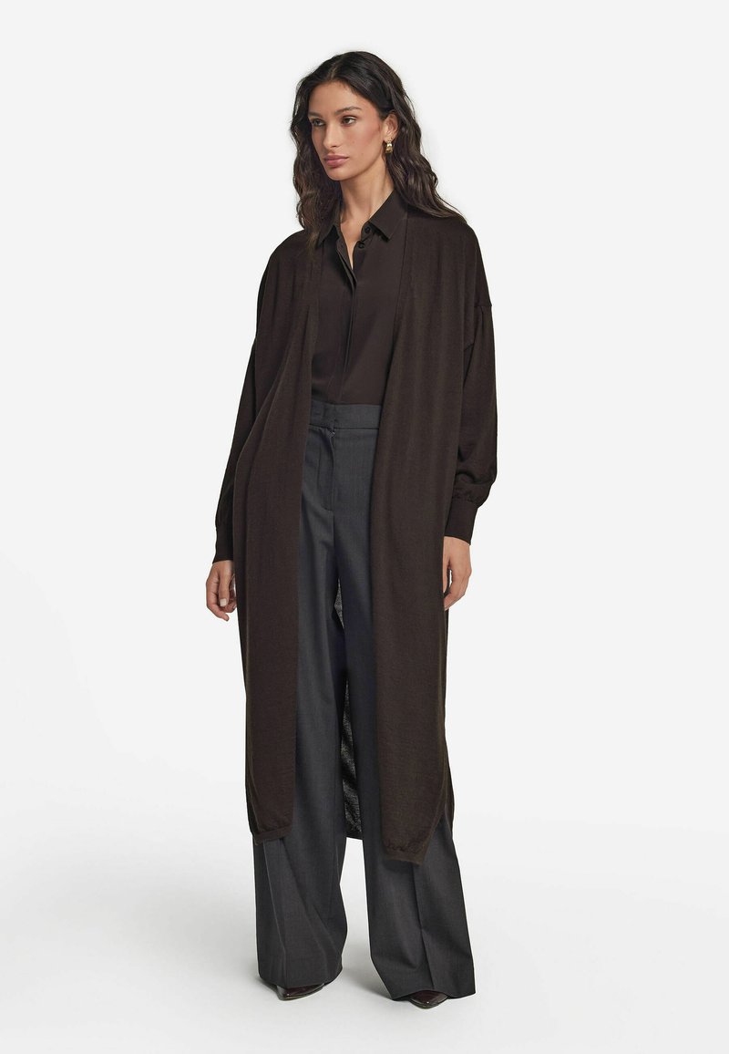 Lång brun kofta i mjukt tyg, med öppen fram och vida ärmar, tillsammans med mörka höga byxor.