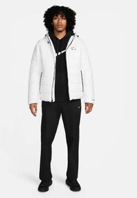 Weißer Puffer-Jacke mit Reißverschluss vorn, getragen über einem schwarzen Hoodie und schwarzen Hosen. Mit Markenlogo auf der Brust und seitlichen Taschen.