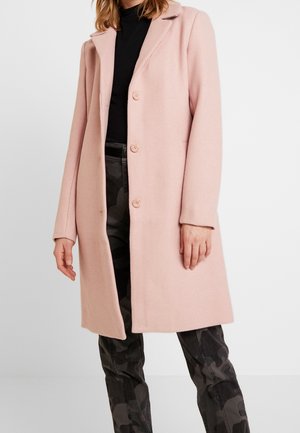 Kobieta ubrana w jasno różowy płaszcz zapinany na guziki, na czarnym golfie i ciemnych spodniach w kamuflażu, na jednolitym białym tle.