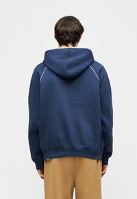 Persoon gekleed in een marineblauwe hoodie met witte bies op de schouders en beige broek, met de rug naar de kijker toe, tegen een eenvoudige lichte achtergrond.