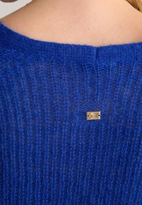 Niebieski sweter dziany z otwartym splotem, z złotą metką z logo przymocowaną z tyłu. Miękka tekstura i swobodny krój.