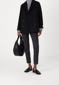 Zwarte dubbelzijdige jas over een gestreepte coltrui, gecombineerd met donkere slim fit jeans en zwarte loafers, terwijl hij/zij een zwarte leren tas vasthoudt.