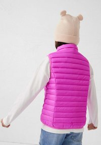 Getailleerd, mouwloos donsjack in felroze met doorgestikte textuur. Draagt over een wit shirt met lange mouwen, gecombineerd met lichtblauwe spijkerbroek en een beige hoed.