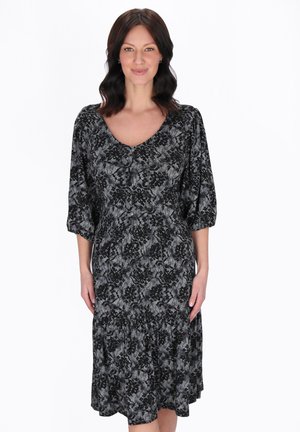 Femeie în picioare, purtând o rochie de lungime până la genunchi, cu model floral negru și gri, cu mâneci trei sferturi, zâmbind pe un fundal alb.