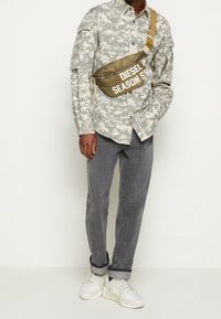 Camouflage-patterned button-up shirt, olive green waist bag with text, gray tapered jeans, and white sneakers with mesh and rubber details.