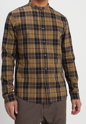 Homme portant une chemise à manches longues boutonnée avec un motif à carreaux jaune moutarde et noir, associée à un pantalon marron.