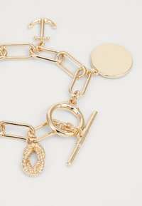 Bracelet en chaîne dorée avec un charme rond, un accent en forme d'ancre et un lien texturé en corde avec un fermoir à bascule. Fini métallique lisse.