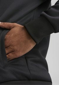 Černá mikina s texturovaným povrchem, obsahující ruku v boční kapse. Manžeta je jednorázová a mírně ribbená.