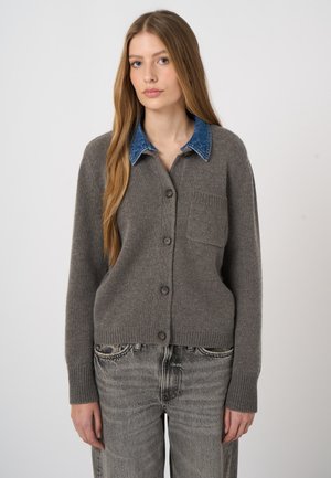 Junge Frau mit langen Haaren, die eine graue, geknöpfte Strickjacke mit blauem Jeanskragen und ausgewaschene graue Jeans vor einem einfarbigen Hintergrund trägt.