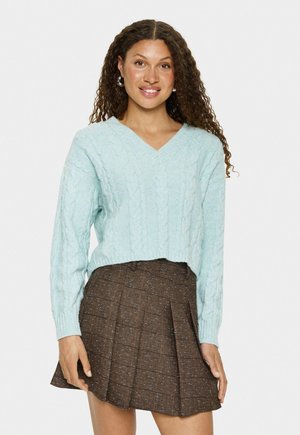 Vrouw met krullend haar, die zilveren creolen draagt, een lichtblauw kabelgebreid cropped sweater en een bruine geplooide minirok.