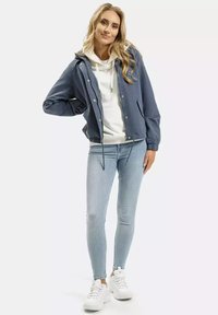 Niebieska kurtka z kapturem zapinana na zatrzaski, noszona na białym hoodie. Połączona z jasnoniebieskimi dopasowanymi dżinsami i białymi sneakersami.