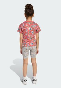 Röd blommig kortärmad t-shirt med gröna och blå accenter, kombinerad med flerfärgade mönstrade cykelbyxor. Vita sneakers med svarta ränder.