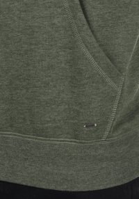 Green sweatshirt with a textured fabric, kangaroo pocket, and metal logo accent near the hem. Features ribbed cuffs and a relaxed fit.