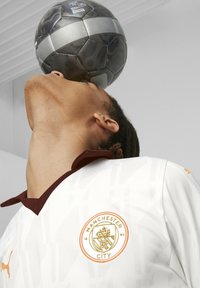 Henkilö, joka pitää yllään valkoista Manchester Cityn pelipaitaa, tasapainottaa mustaa ja harmaata jalkapalloa leuallaan yksinkertaisen taustan edessä.