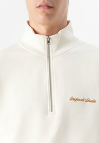 Et close-up af en persons overkrop iført en cremet zip-up sweater med en lille "Original Studio" stiksyning på brystet.