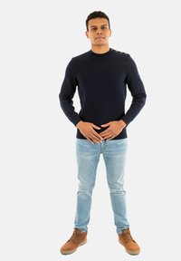 Pullover blu navy con scollatura a girocollo e dettaglio con bottoni sulle spalle, abbinato a jeans azzurri e scarpe marroni stringate, in piedi su uno sfondo bianco.