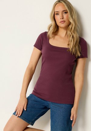 Vrouw met lang blond haar, gekleed in een aansluitend paarse korte mouw top en blauwe spijkershort, staand tegen een lichte muur.