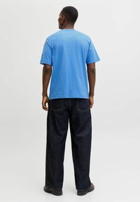 Sinine puuvillane T-särk lühikeste varrukatega, ümar kaelus, kombineeritud tumedate teksapükste ja mustade kingadega, vaadatuna seljalt.