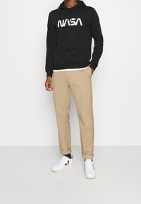 Schwarzer Kapuzenpullover mit weißem "NASA"-Logo, kombiniert mit beigen, schmal zulaufenden Hosen und weißen Turnschuhen mit schwarzen Akzenten.