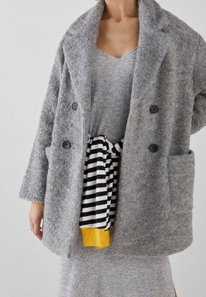 Manteau croisé en mélange de laine gris avec de grands boutons et deux poches à l'avant, porté sur une robe grise avec une cravate rayée et des bordures jaunes.