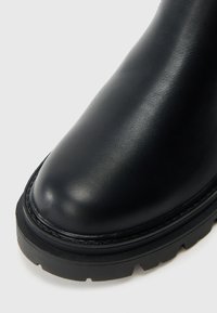 Μαύρο δερμάτινο μποτάκι αστραγάλου με στρογγυλεμένη μύτη, υφή επιφάνειας και παχιά λαστιχένια σόλα με έντονα ανάγλυφα πέλματα. Ορατή ενισχυμένη ραφή.