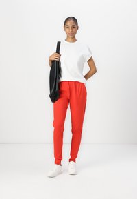 Röda avsmalnande byxor med elastisk midja, i kombination med en vit t-shirt med kort ärm och vita sneakers. Håller en svart tote-väska.