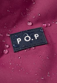 Lähikuva vesipisaroista violetilla kankaalla, jossa on musta suorakulmainen etiketti, jossa lukee "P.O.P" valkoisilla kirjaimilla.