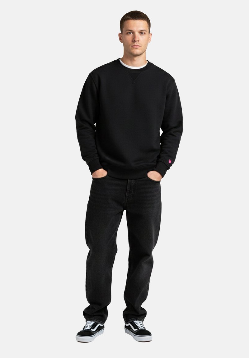 Jeune homme debout, les mains dans les poches, portant un sweat-shirt noir, un jean noir et des baskets noir et blanc sur un fond uni.