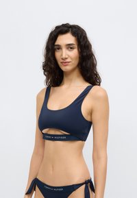 Σετ μπικίνι σε ναυτικό μπλε με σχέδιο κοψίματος. Το πάνω μέρος έχει πλατιά τιράντες και λαστιχένιο κάτω μέρος με λογότυπο. Το κάτω μέρος διαθέτει λεπτομέρειες με κορδόνια στα πλαϊνά. Ομαλή υφή.