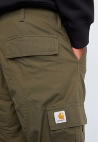 Pantalon cargo vert olive avec un tissu texturé, deux poches arrière et un patch logo Carhartt sur la poche latérale.