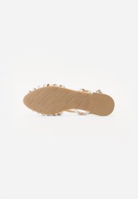 Sandale plate avec une semelle beige, des brides argentées tressées, et de petits gemmes transparentes le long du bord. Semelle en caoutchouc mat texturé avec une marque.