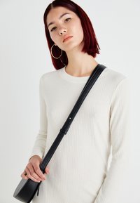 Camisola de manga longa com textura em off-white, acompanhada de uma mala tiracolo preta com uma alça fina. A modelo tem brincos de argola e cabelo vermelho.