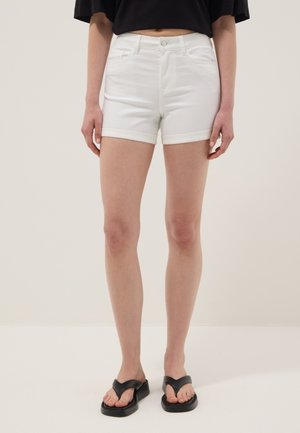 Vrouw draagt witte omgeslagen denimshorts, zwarte cropped top en zwarte platform slippers, staand op een effen lichte achtergrond.