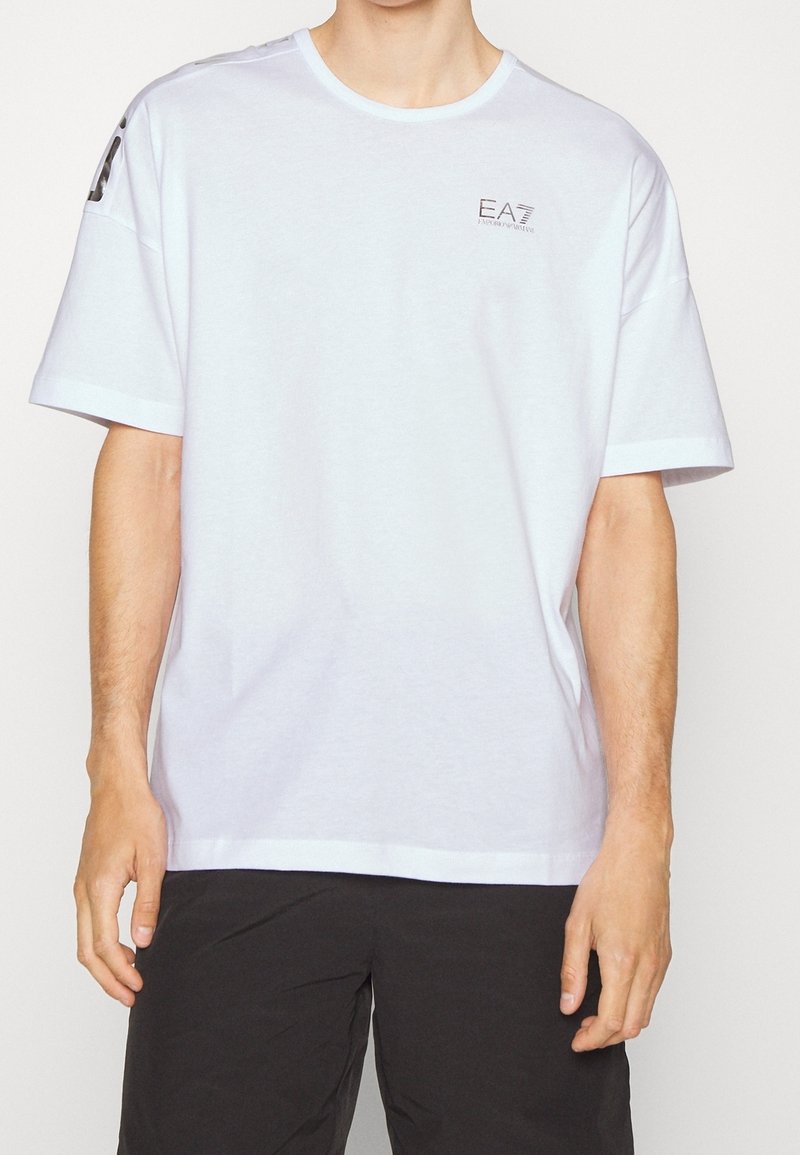 Camiseta blanca de manga corta hecha de algodón, con escote redondo y logo EA7 en el lado izquierdo del pecho, combinada con pantalones cortos oscuros.