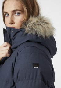 Námornická modrá zimní bunda se zipem vpředu, kapucí s kožešinovým lemem a černou nášivkou na levém rameni. Texturovaná látka.